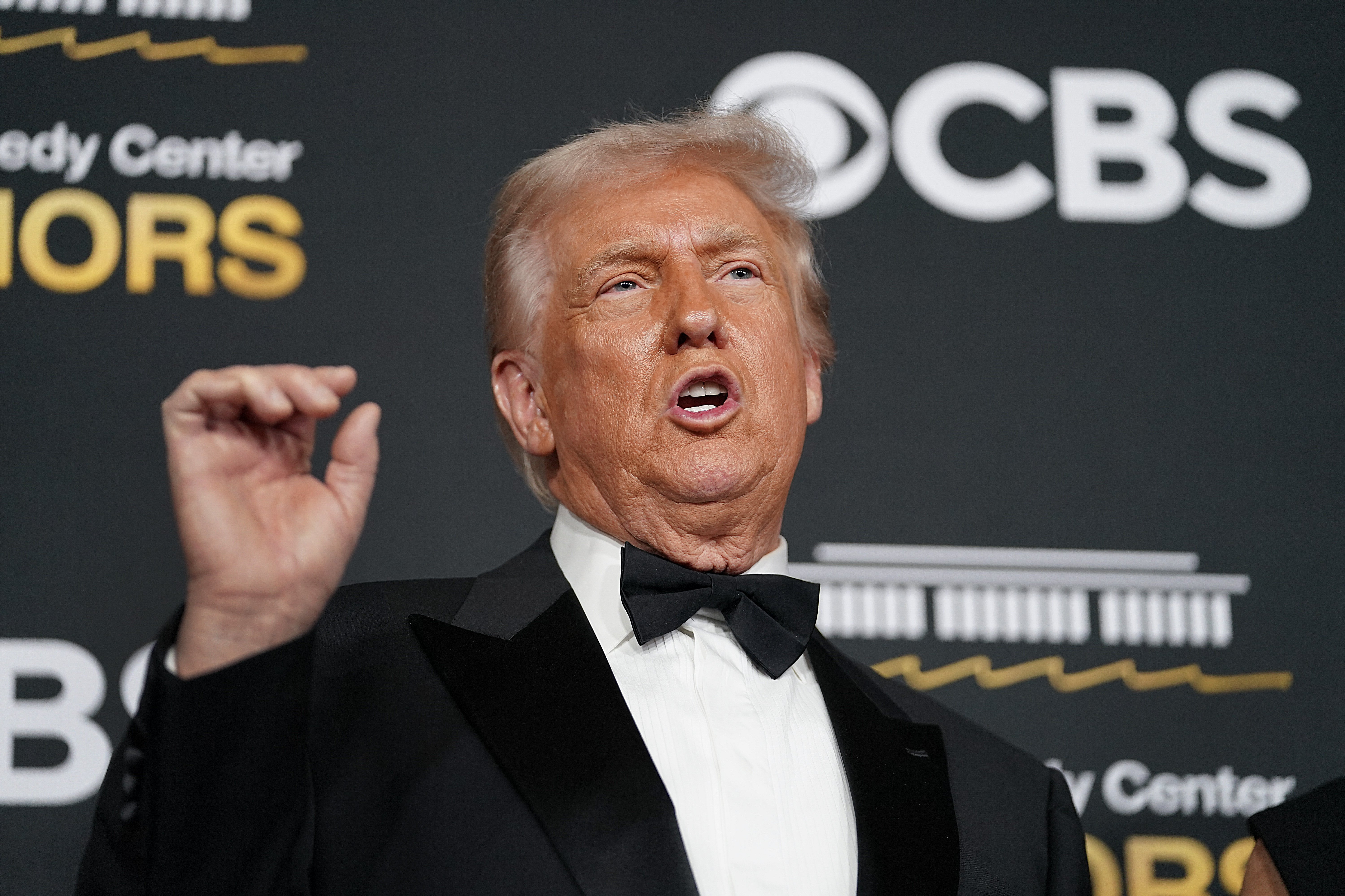 An older man with light hair, wearing a black tuxedo and bow tie, gestures with his raised right hand in front of a backdrop displaying the CBS logo and "Kennedy Center Honors" text.