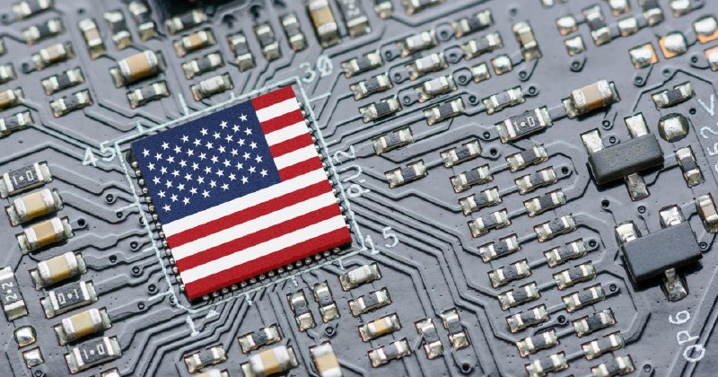 A square chip displaying the United States flag is mounted on a densely populated circuit board surrounded by small electronic components.