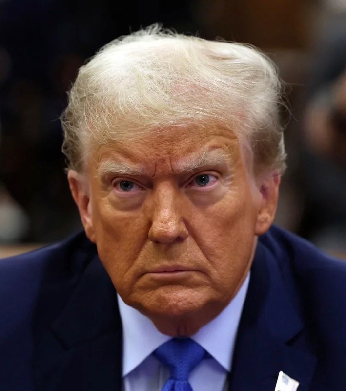A close-up photograph shows an older man with light blonde hair, wearing a dark suit, light blue shirt, and blue tie, looking directly forward with a serious expression.