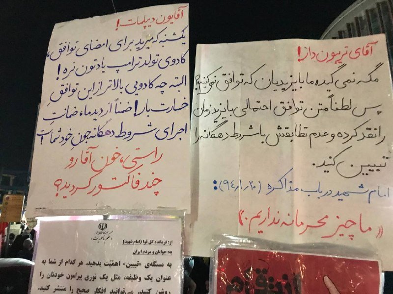 Two handwritten Persian protest signs in red and blue ink are displayed above an official Iranian government notice at night.