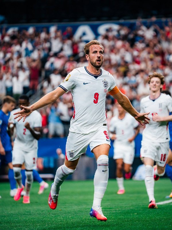 A soccer player wearing a white jersey with the number 9 runs with arms outstretched in celebration on a green pitch, surrounded by teammates and a stadium crowd.