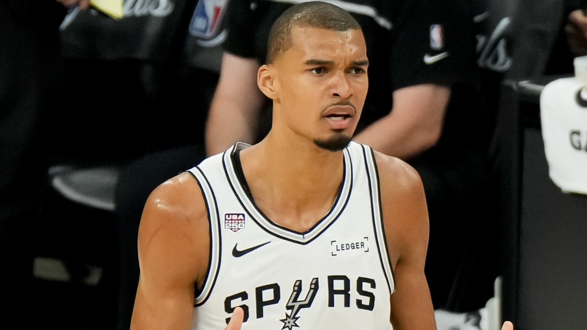 A basketball player wearing a white San Antonio Spurs jersey sits on the bench during a game, with a USA 250 patch and Ledger sponsor logo visible.