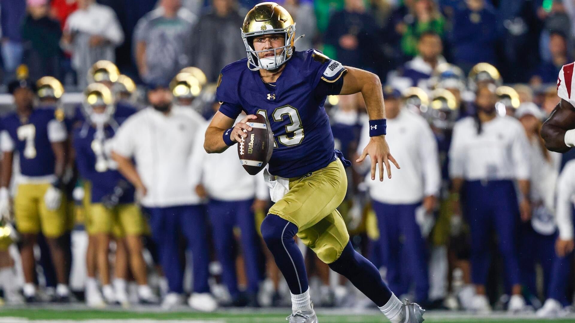A football player wearing a navy blue jersey with the number 13 and a gold helmet runs with the football, with teammates and coaches visible on the sideline behind him.