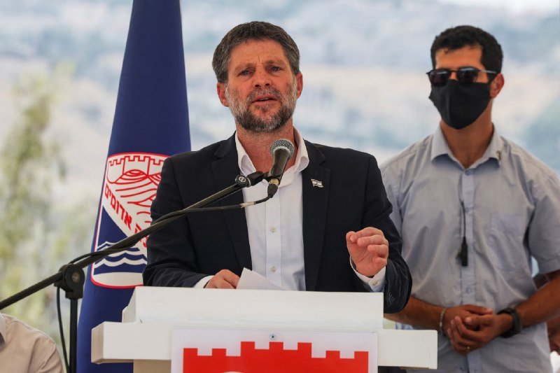 A bearded man in a dark blazer speaks at a podium bearing a red city emblem, gesturing with one hand, while a masked man stands behind him near a blue flag.