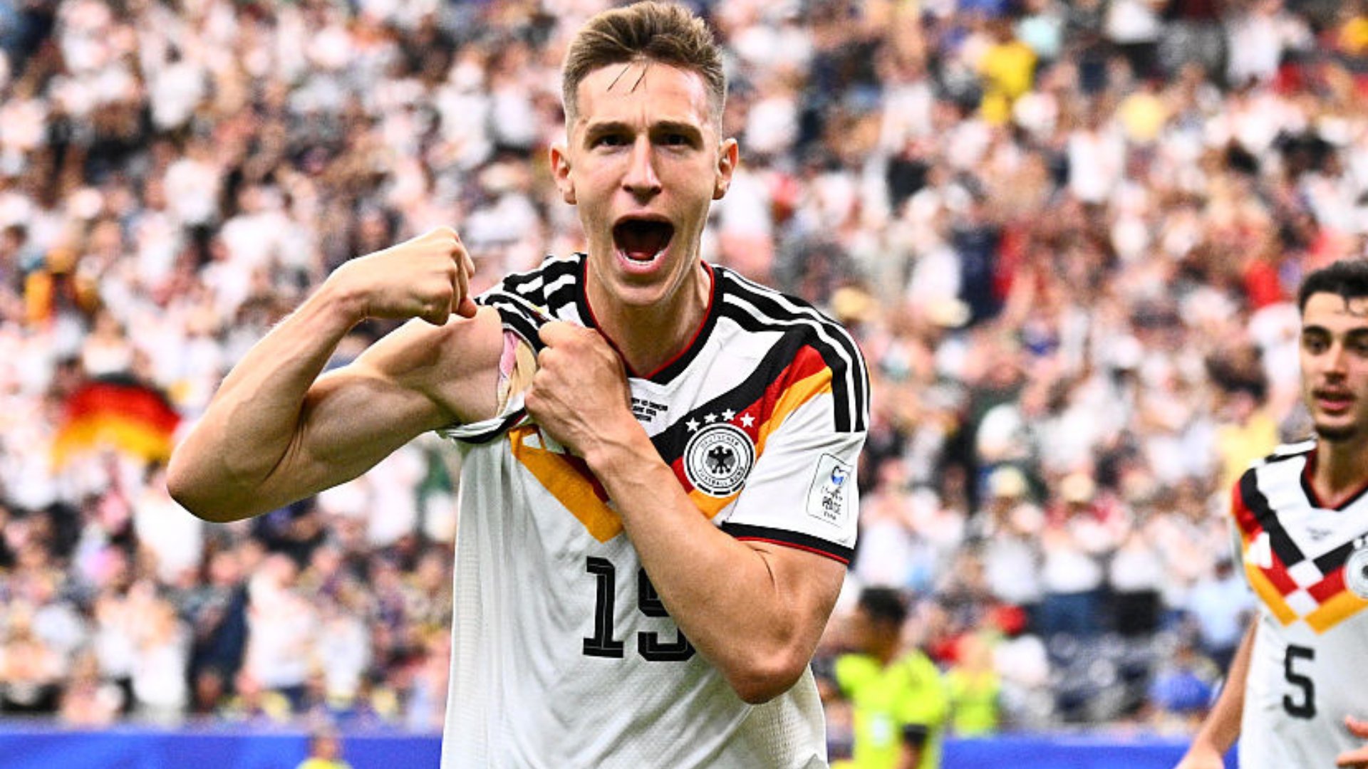 A soccer player in a white Germany jersey (#13) celebrates with clenched fists and an open-mouthed shout in front of a stadium crowd, while a teammate (#5) stands nearby.