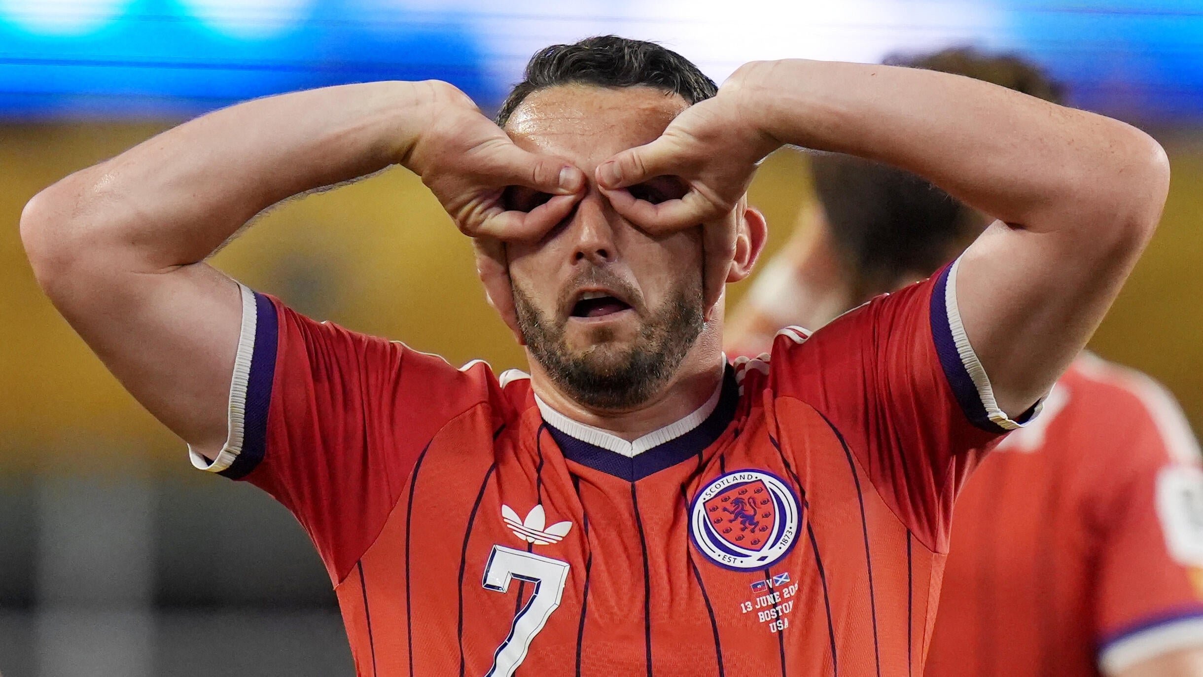 A soccer player wearing a red Scotland jersey with the number 7 covers his eyes with both hands, appearing visibly emotional.