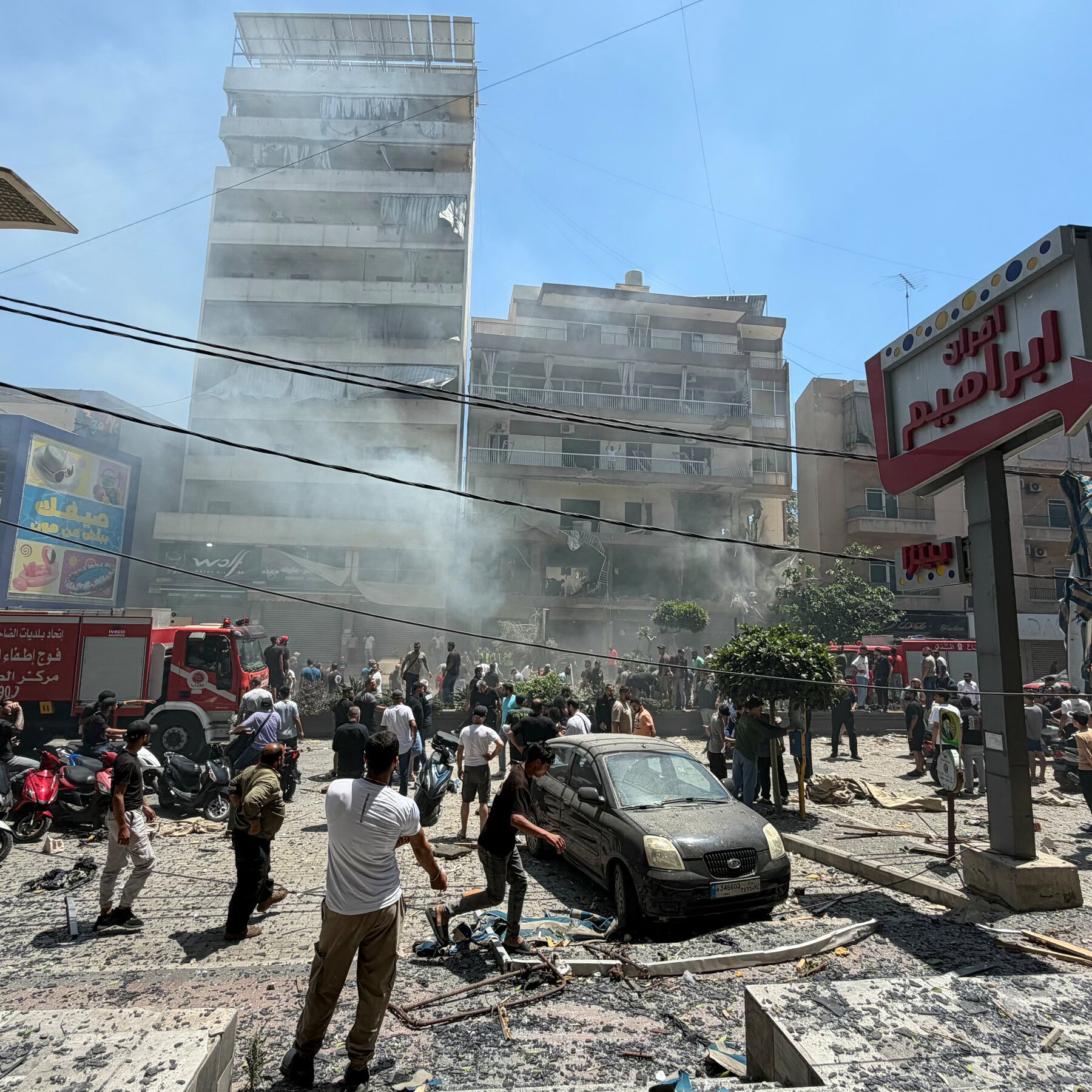 Crowds gather in a debris-strewn street outside smoke-damaged buildings, with a red fire truck and a dust-covered damaged car visible amid scattered rubble.