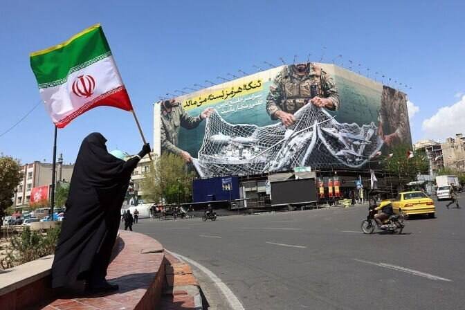 A woman in a black chador holds an Iranian flag while standing near a large billboard depicting military personnel pulling a net over a US aircraft.