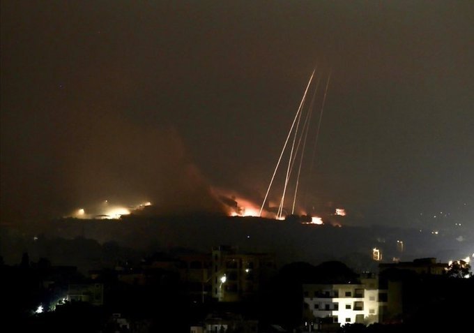 Bright streaks arc through a night sky above a hillside lit by flames and smoke, with silhouetted buildings visible in the foreground.