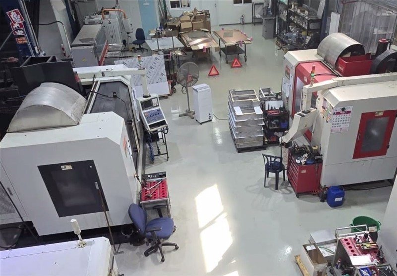 An empty industrial workshop contains large white and red CNC machines, tool carts, a pedestal fan, office chairs, and metal storage bins on a gray floor.
