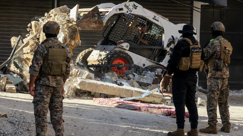 Three armed personnel in military gear and tactical vests stand amid rubble near a damaged Bobcat skid-steer loader and debris outside a closed storefront.