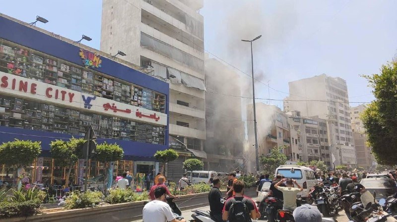 Thick black smoke rises from a damaged building behind a storefront labeled "SINE CITY," as bystanders gather on the street with parked motorcycles and vehicles nearby.
