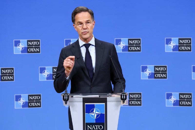 A man in a dark suit and tie stands at a podium with the NATO logo, speaking and gesturing with his right hand against a blue backdrop displaying repeated NATO/OTAN logos.