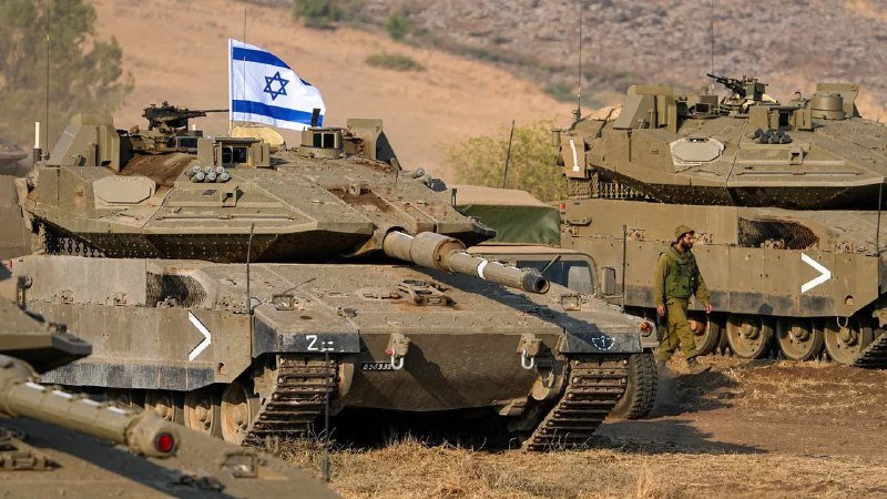 Israeli flag flies atop a Merkava tank as a soldier in green uniform walks past armored military vehicles parked in a dry, hilly field.