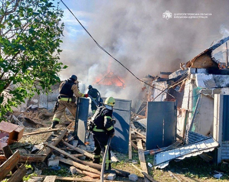 Firefighters in helmets and protective gear spray water on flames engulfing a collapsed building amid scattered debris and smoke, with a Cyrillic military administration watermark in the corner.