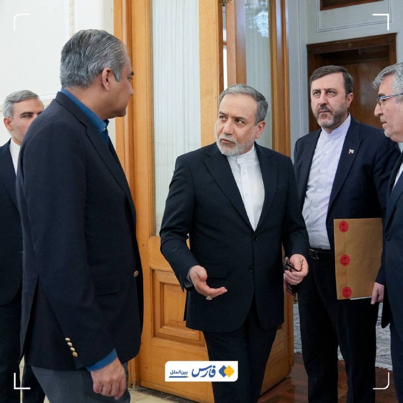 Five men in suits stand in a formal indoor setting near wooden doors, with one bearded man gesturing while speaking to the others.