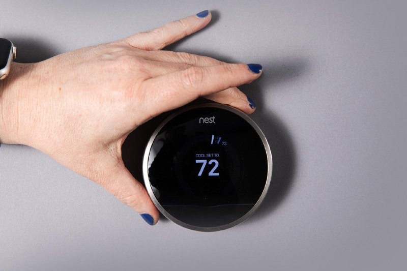 A hand with blue nail polish rests on a black circular Nest thermostat mounted against a gray wall, displaying a set temperature of 72 degrees in "COOL SET TO" mode.
