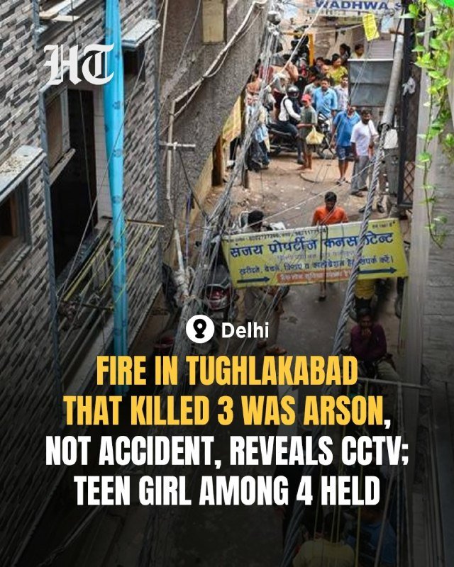 A crowded narrow alleyway in Delhi shows people gathered near buildings with overhead wires and Hindi signage.