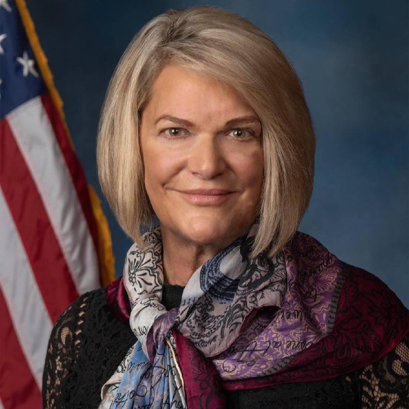 A woman with shoulder-length blonde hair smiles at the camera, wearing a black top and patterned scarf, with an American flag and blue backdrop behind her.