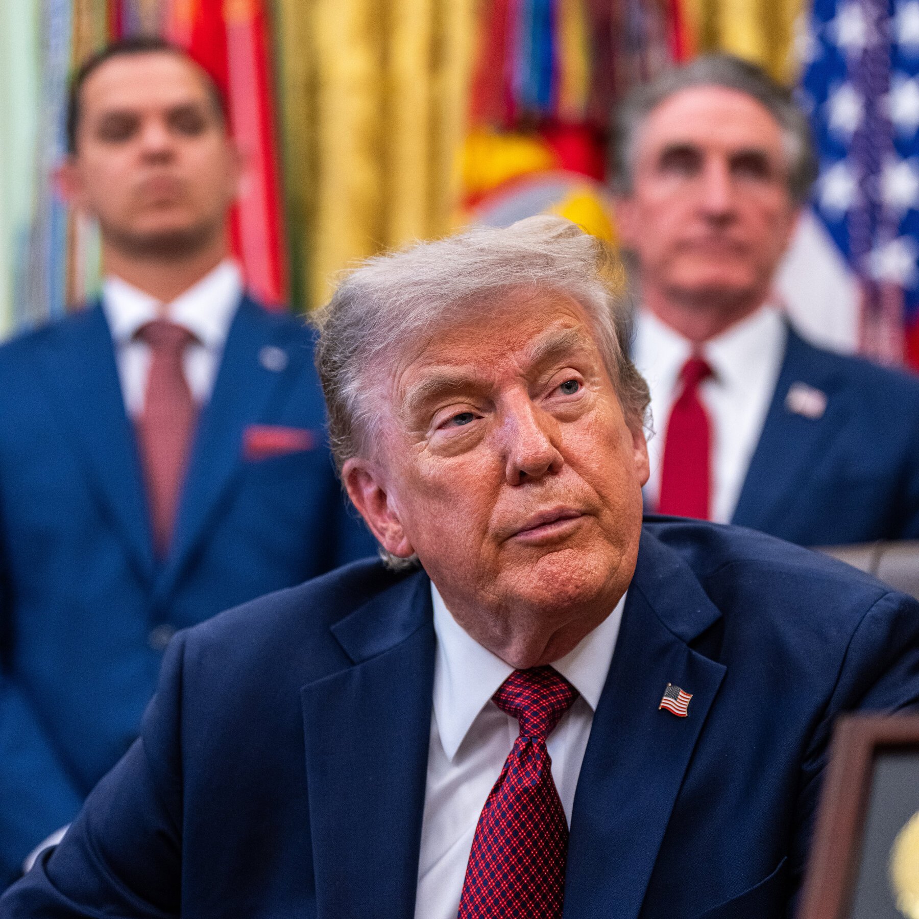 A seated older man with white hair, wearing a navy suit with an American flag pin and red patterned tie, looks upward, with two other men in suits and a US flag visible behind him.