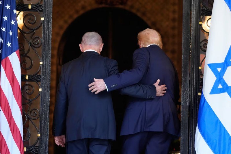 Two men in dark suits walk arm-in-arm through an ornate doorway, flanked by an American flag on the left and an Israeli flag on the right.