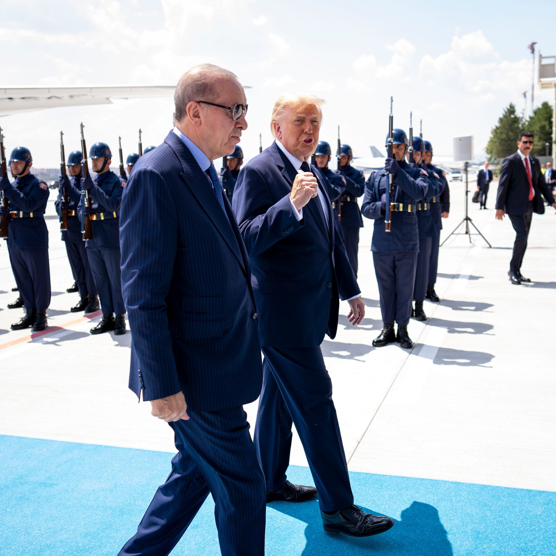 Two men in dark suits walk along a blue carpet on a tarmac, passing a military honor guard in dark blue uniforms and helmets holding rifles.