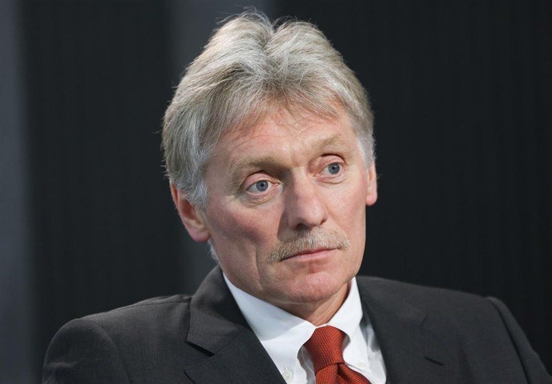 A gray-haired man with a mustache, wearing a dark suit and red tie, looks off-camera against a dark background.