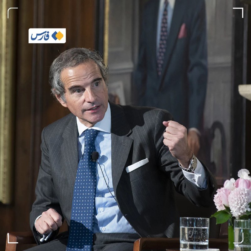 A gray-haired man in a dark suit and blue dotted tie gestures with his hand while speaking, with a standing figure, a glass of water, and pink flowers nearby.
