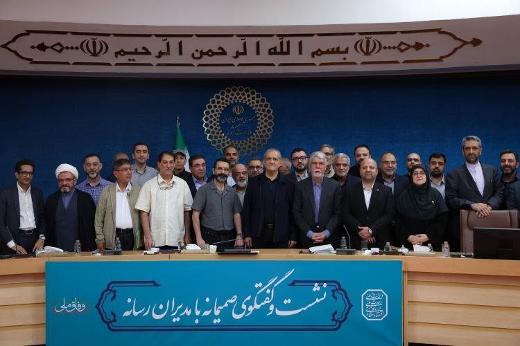 A large group of men and two women pose for a photo behind a long desk with a teal banner bearing Persian script, beneath an emblem and Islamic calligraphy.