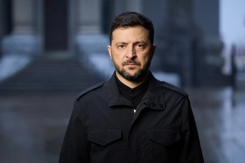 A bearded man in a black jacket looks directly at the camera in front of an out-of-focus gray stone building.
