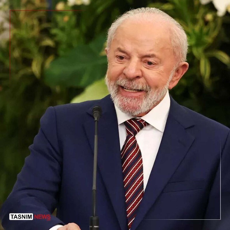 A white-bearded man in a navy blue suit and striped tie smiles while speaking at a microphone, with green plants visible in the background.