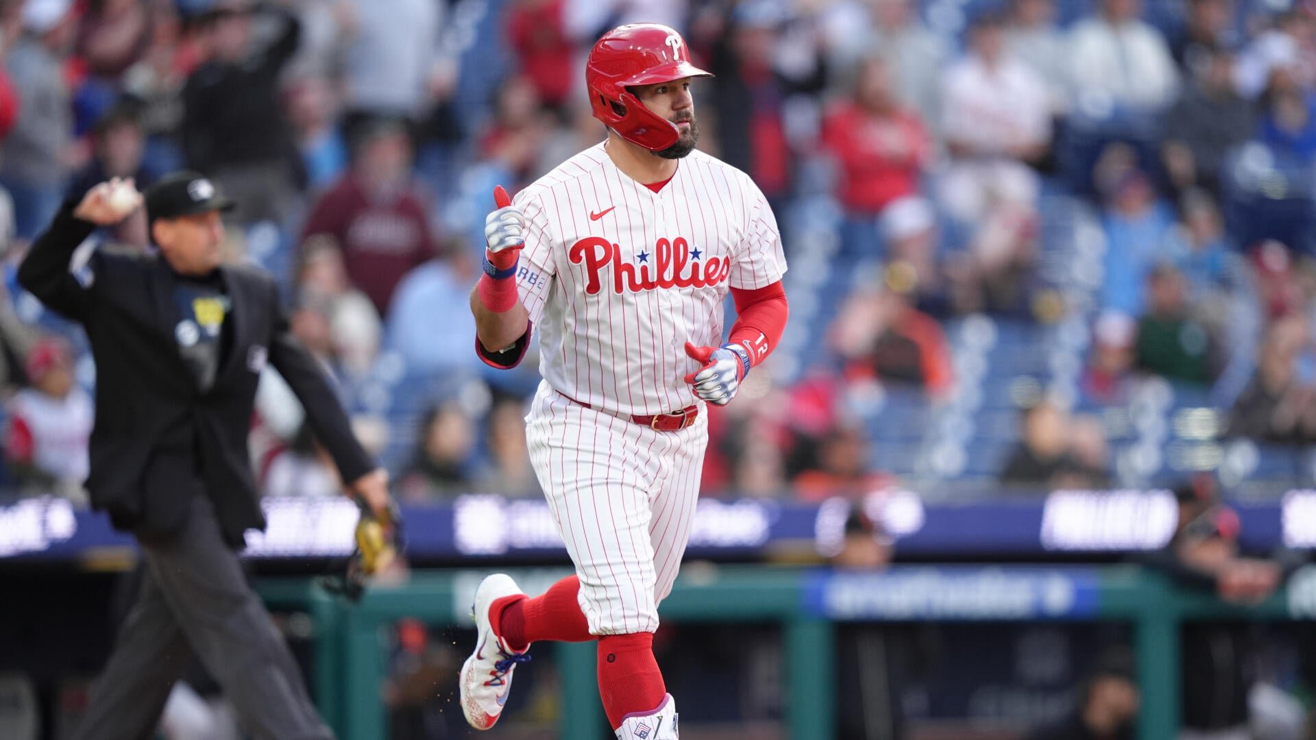 A Philadelphia Phillies player in a red helmet and pinstriped uniform runs the bases, giving a thumbs-up gesture, as an umpire watches from the background.