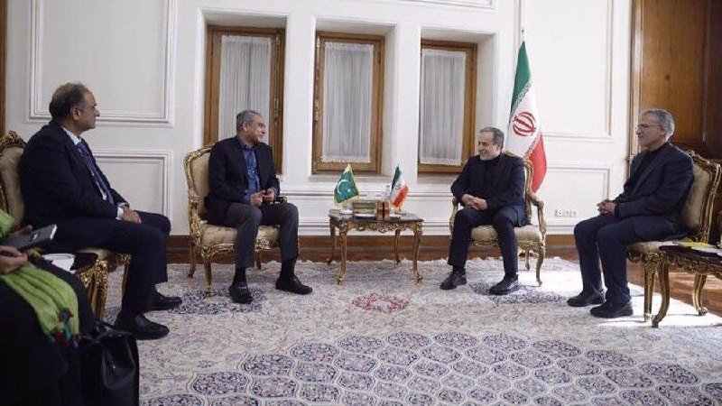Four men in dark suits sit facing each other on ornate chairs in a formal room, with a large Iranian flag and smaller table flags visible behind them.