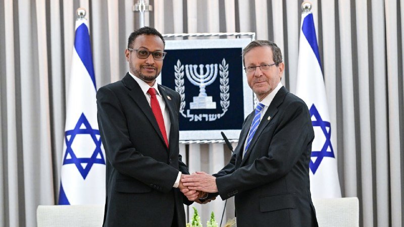 Two men in dark suits shake hands in front of Israeli flags and the official emblem of the State of Israel.