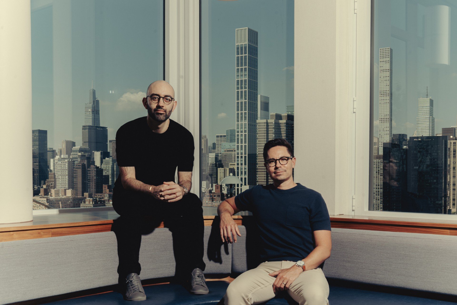 Two men wearing glasses sit on a gray bench in a high-rise office with floor-to-ceiling windows overlooking a city skyline.