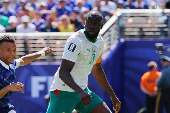 Two soccer players, one in a white jersey and one in blue, compete during a match in a stadium filled with spectators.