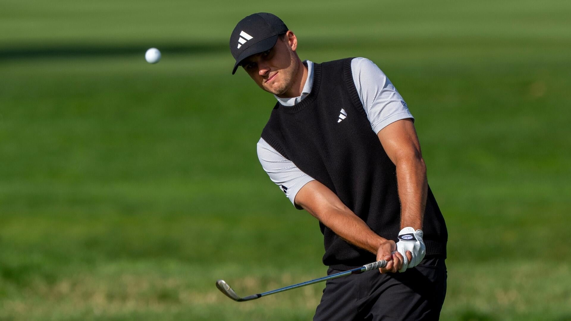 A golfer in a black vest and cap hits a chip shot onto a green, the ball visible in mid-air beside him.
