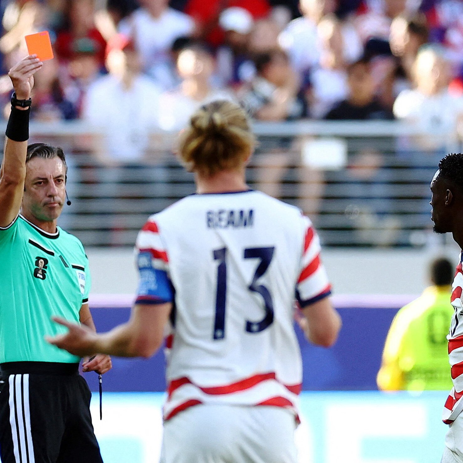 A referee in a green jersey raises a red card toward a soccer player wearing a white USMNT jersey with "REAM" and the number 13.