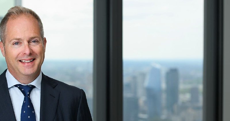 A smiling man in a dark suit and patterned blue tie stands indoors near a large window with a city skyline visible in the background.