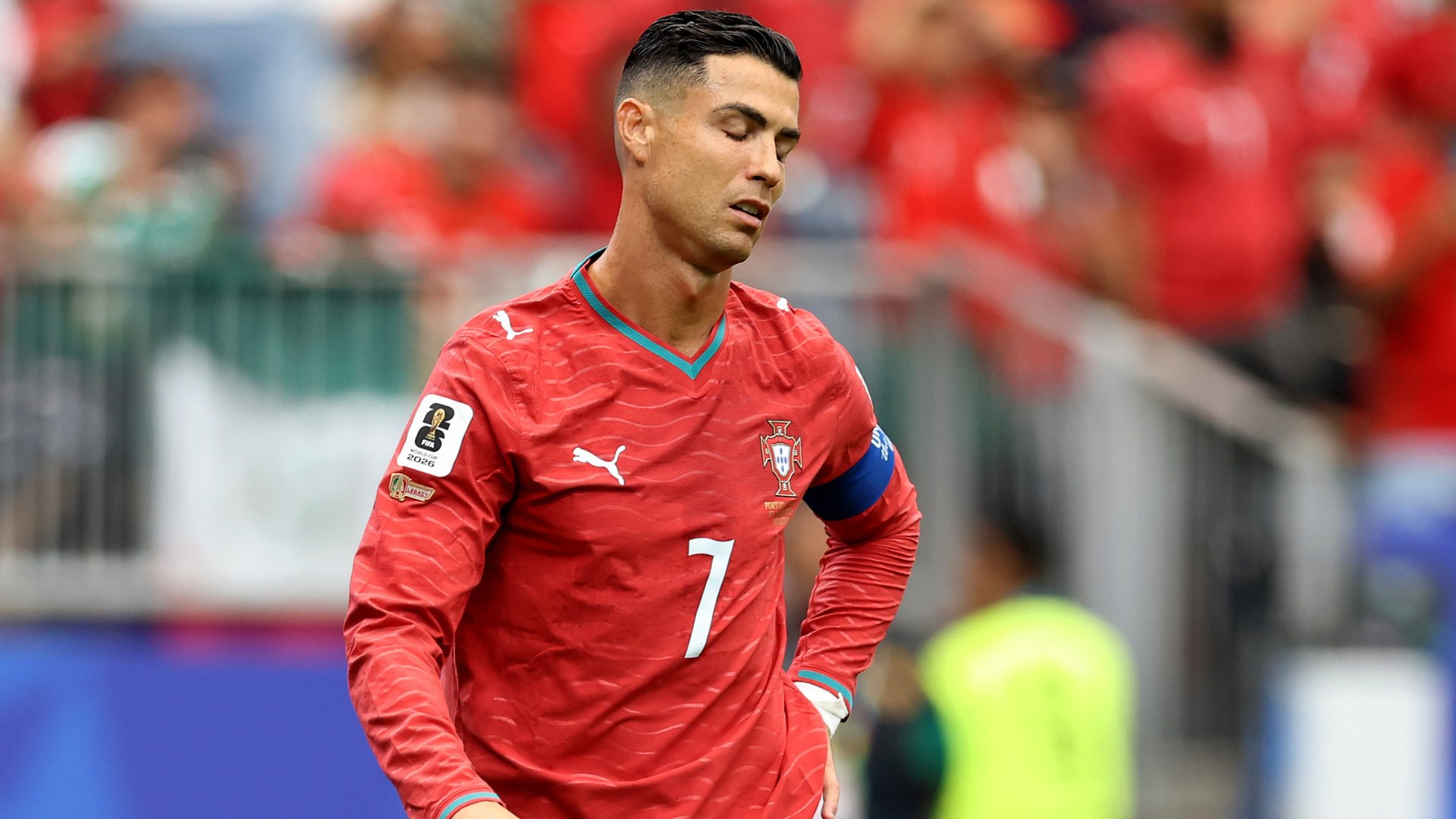 A soccer player wearing a red jersey with the number 7 stands on a pitch with blurred red-clad fans visible behind him.