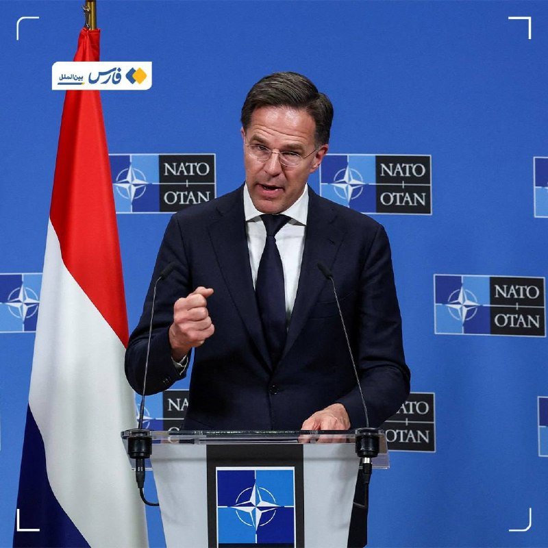 A man in a dark suit and tie speaks at a NATO-branded podium, gesturing with one hand, against a blue backdrop featuring NATO/OTAN logos and a Dutch flag.