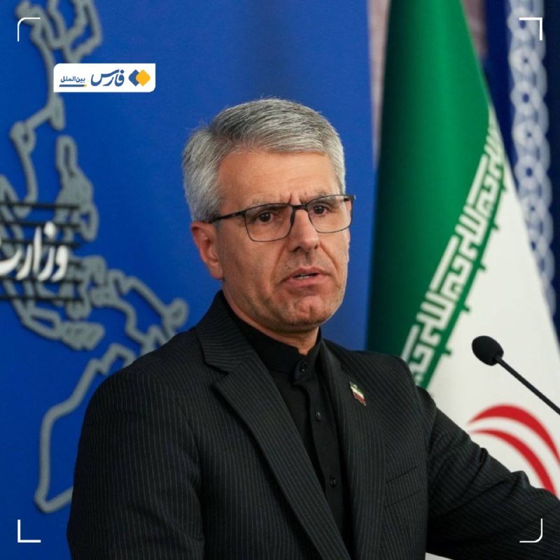 A gray-haired man wearing glasses and a dark pinstripe suit speaks at a podium with a microphone, positioned in front of an Iranian flag and a blue backdrop featuring a map outline.