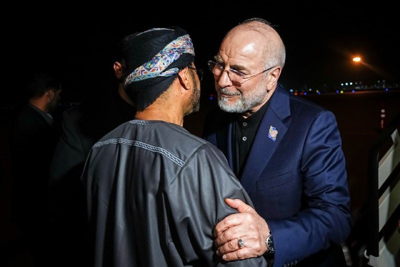 A bearded man in a navy suit and glasses warmly embraces a man wearing a patterned headband and gray robe on an airport tarmac at night.
