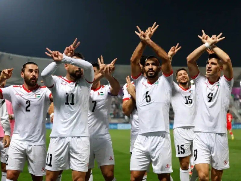 A group of soccer players in white uniforms with red and green accents celebrate on the field, raising their arms with fingers forming V-signs.