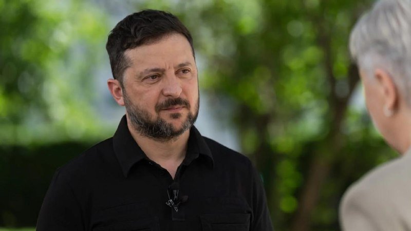 A bearded man in a black shirt listens attentively to a woman with short gray hair during an outdoor interview setting with green foliage in the background.