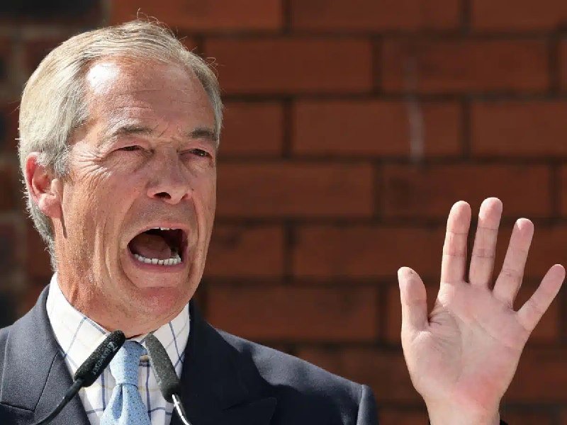 Nigel Farage speaking animatedly with raised hand and open mouth at a podium microphone against a brick wall background.