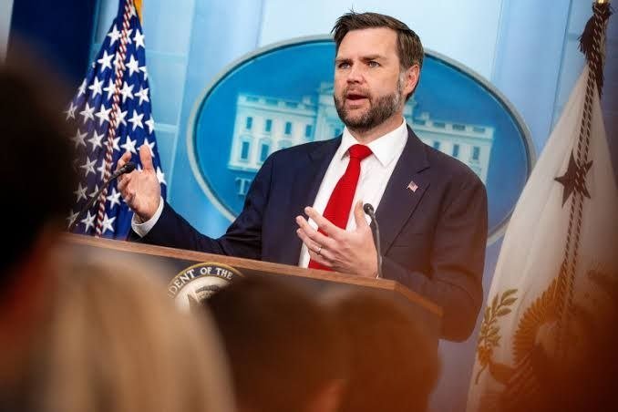 A bearded man in a dark suit and red tie speaks at a podium bearing a presidential seal, gesturing with both hands in front of a White House backdrop and flags.