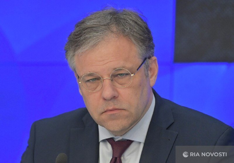 A gray-haired man wearing round glasses, a dark suit, and a maroon tie sits before a blue background, with "RIA NOVOSTI" watermarked in the corner.