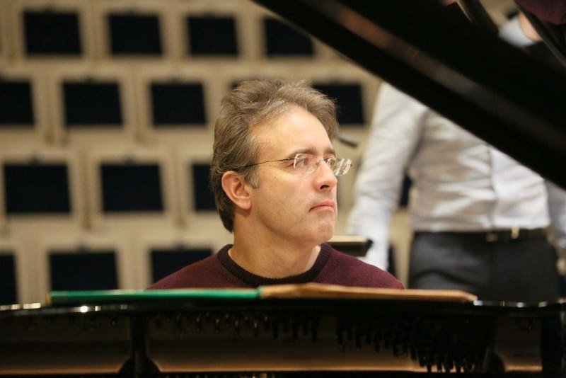 A bespectacled man in a maroon sweater sits at a grand piano, with another person in a white shirt standing behind him in a paneled room.
