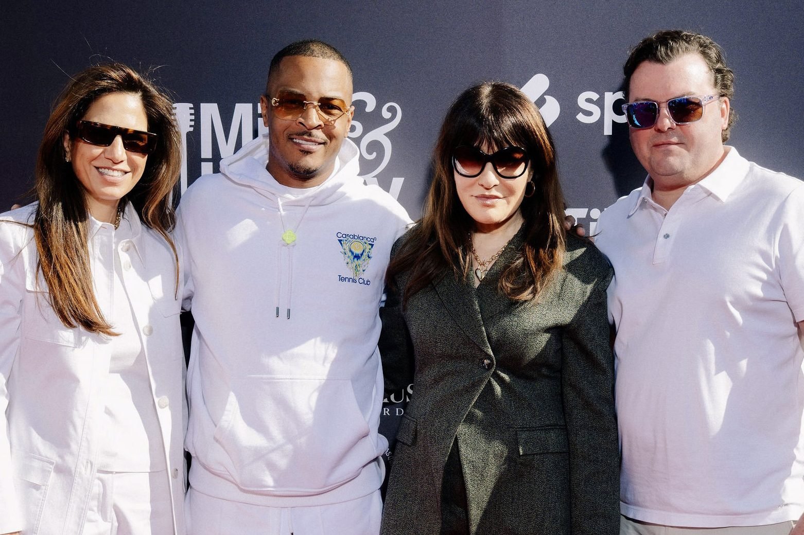 Four people pose together in front of a dark backdrop featuring partial white lettering and logos, with three wearing sunglasses and the man in a white hoodie reading "Casablanca Tennis Club."